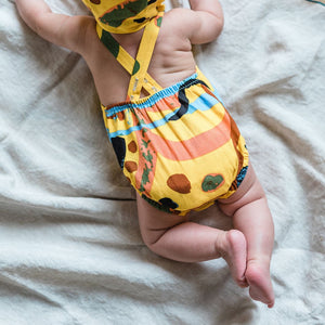 Baby lying on tummy on blanket in bright printed romper and bonnet for Petits Genoux